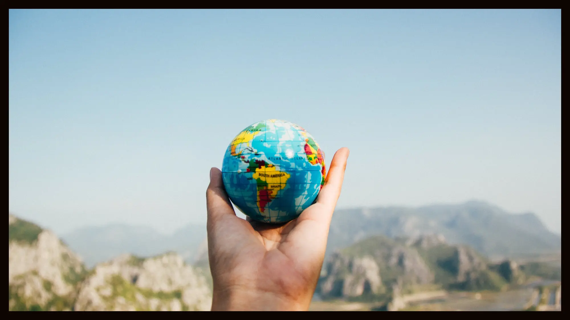A hand holding a small globe with a blurry mountainous landscape in the background, symbolizing travel and the beauty of exploring the world.
