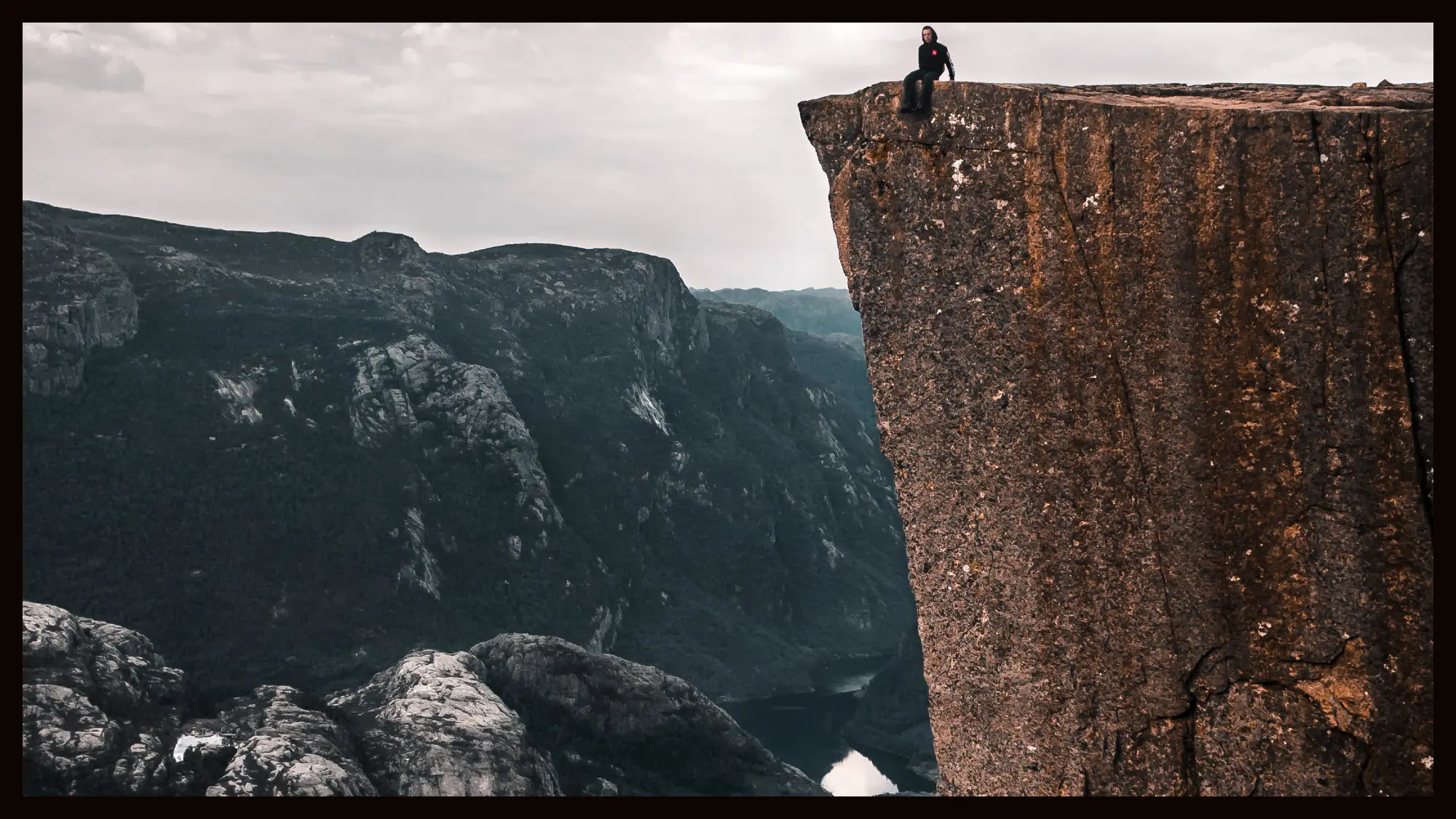 A solitary figure standing on the edge of a high cliff overlooking a deep valley with rugged mountains in the background, conveying a sense of adventure and solitude.