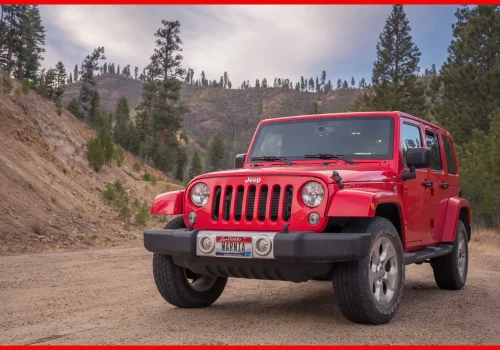 A red Jeep Wrangler parked on a dirt road with a forested mountainous backdrop under a cloudy sky.