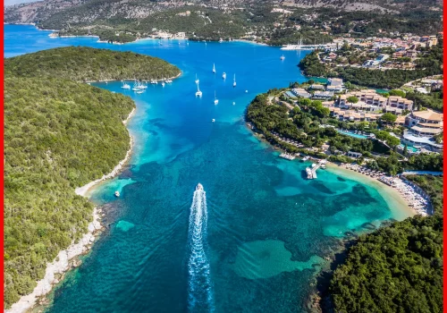 An overhead view of a yacht creating a white wake in the azure waters of a bay, surrounded by greenery and coastal resorts.