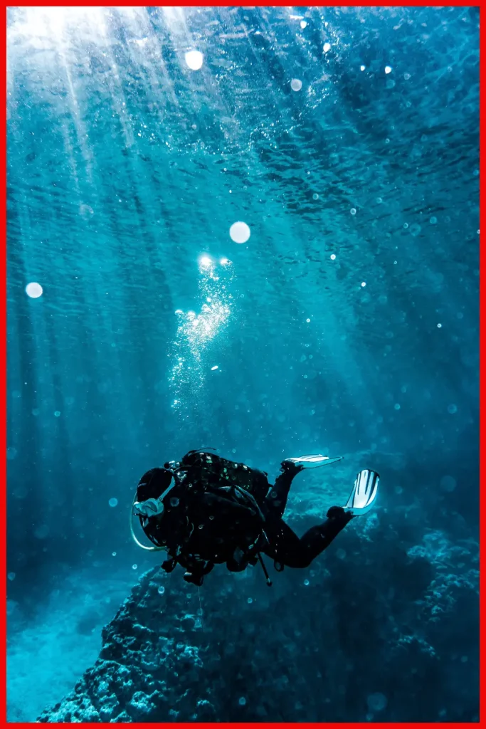 A diver in black wetsuit swims near the ocean floor, surrounded by bubbles and the deep blue sea, highlighting the isolation of the underwater world.